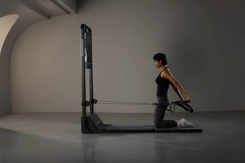 A woman kneeling on a fitness mat, using a resistance machine with cables, while performing a stretching exercise.