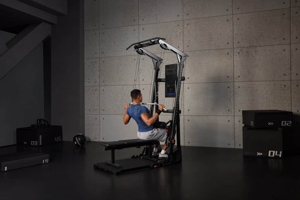 A person performing a seated exercise on a versatile gym equipment, focusing on strength training in a modern gym setting with concrete walls.