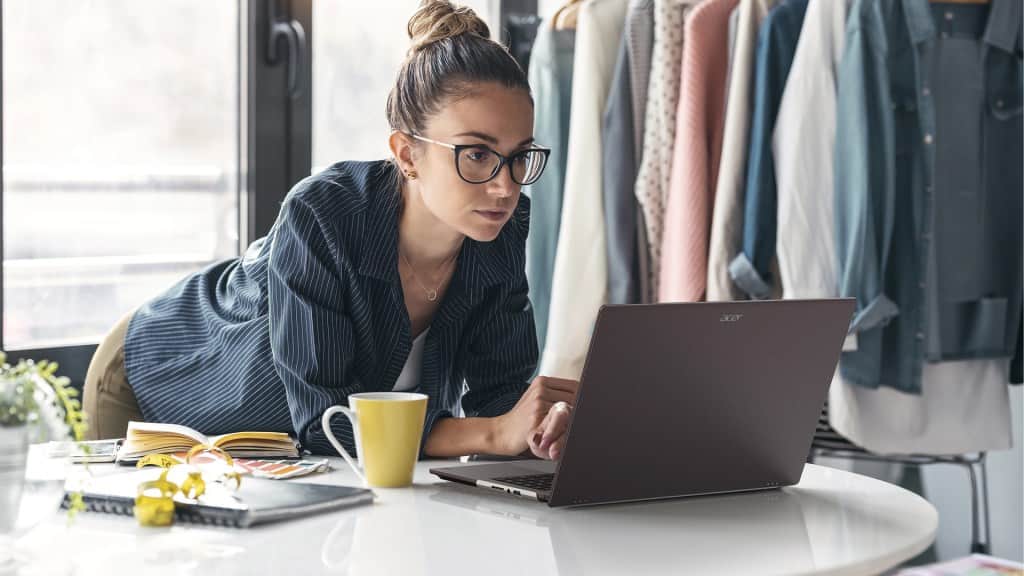 A focused woman wearing glasses works on a laptop at a table surrounded by colorful clothing and stationery.