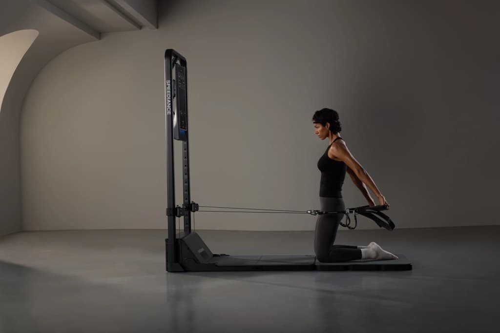 A woman kneeling on a fitness mat, using a resistance machine with cables, while performing a stretching exercise.