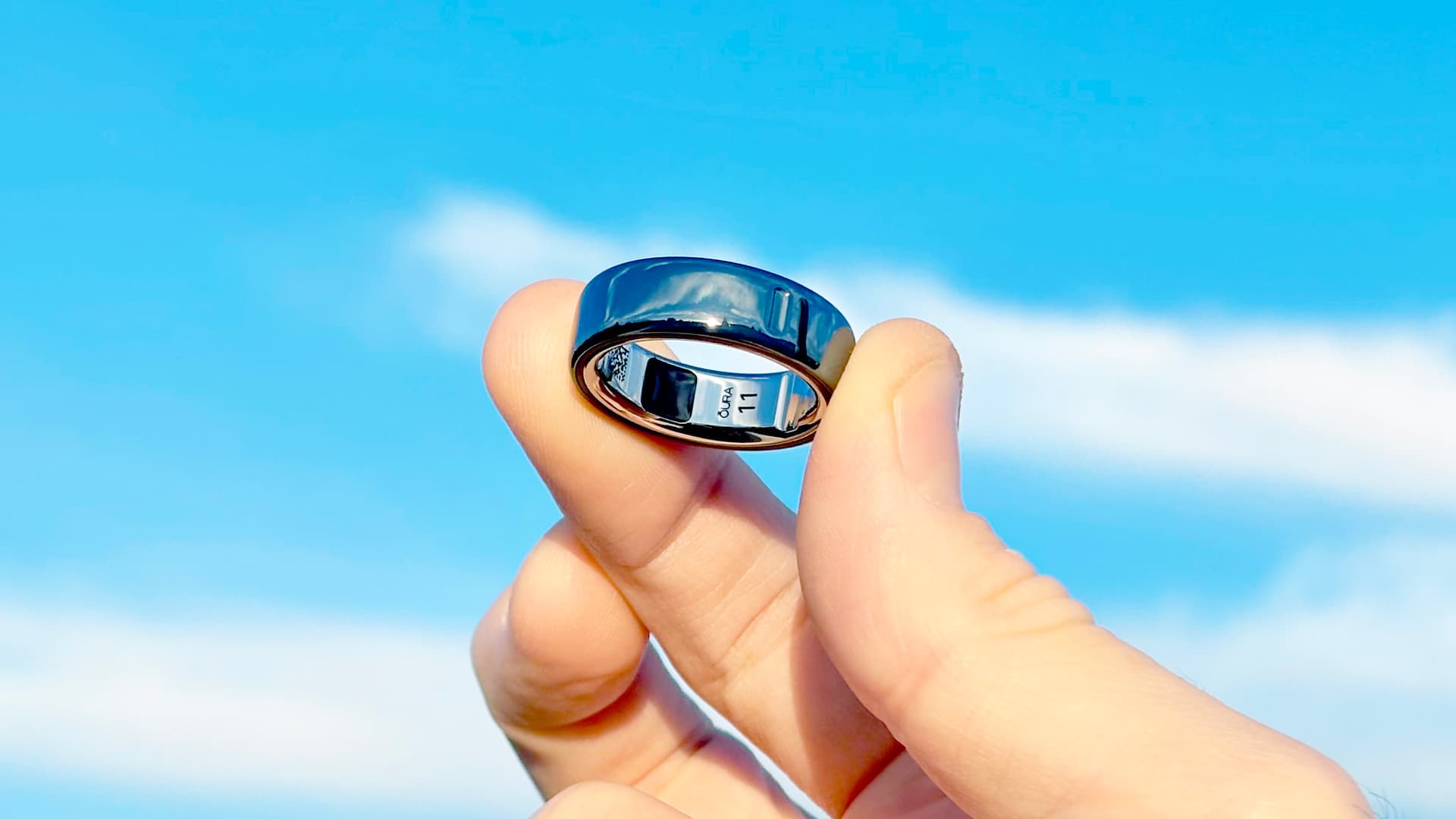 Close-up of the Oura Ring 4 shining in sunlight in a user's hand, with blue skies and clouds behind it — Image credit: Dan Bracaglia
