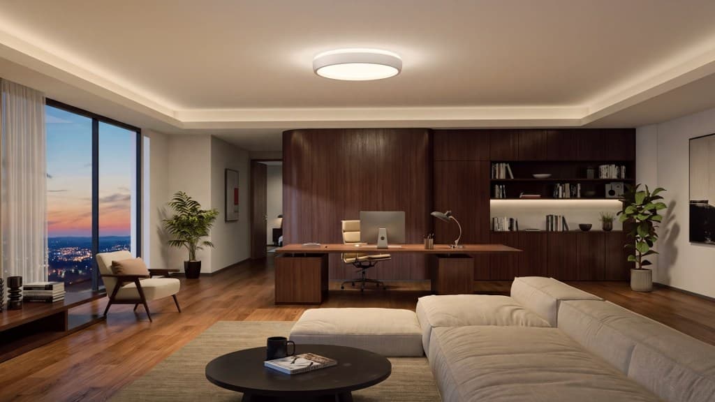 A modern office interior featuring a wooden desk with a computer, ergonomic chair, and desk lamp. The space includes a comfortable beige sectional sofa, a round coffee table, and large windows with a city view at sunset. Decorative plants and bookshelves add a touch of elegance.
